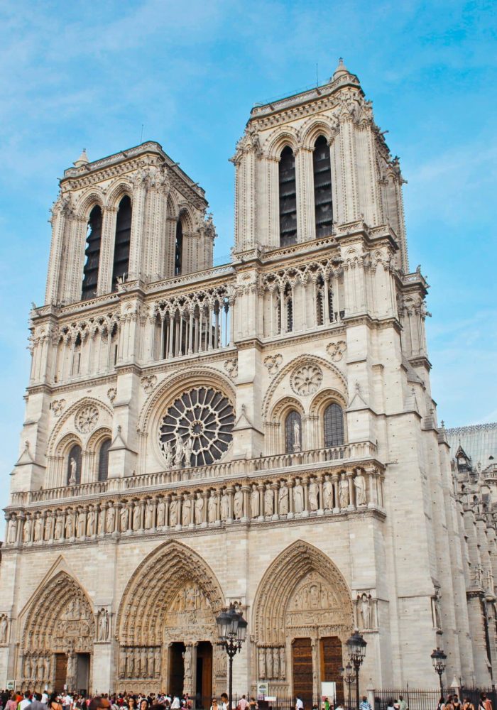 Notre-Dame de Paris cathedral, with its iconic twin towers, ornate façade and large rose window, stands out against a blue sky as crowds gather at its entrance.