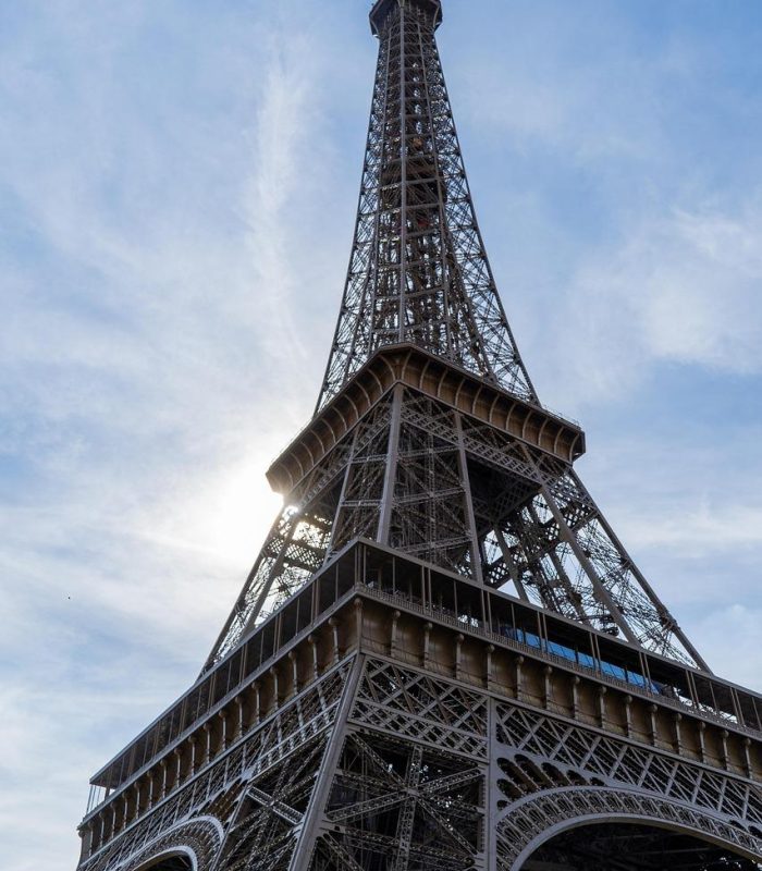 Vue en contre-plongée de la Tour Eiffel dans un ciel bleu partiellement nuageux, la lumière du soleil traversant la structure.