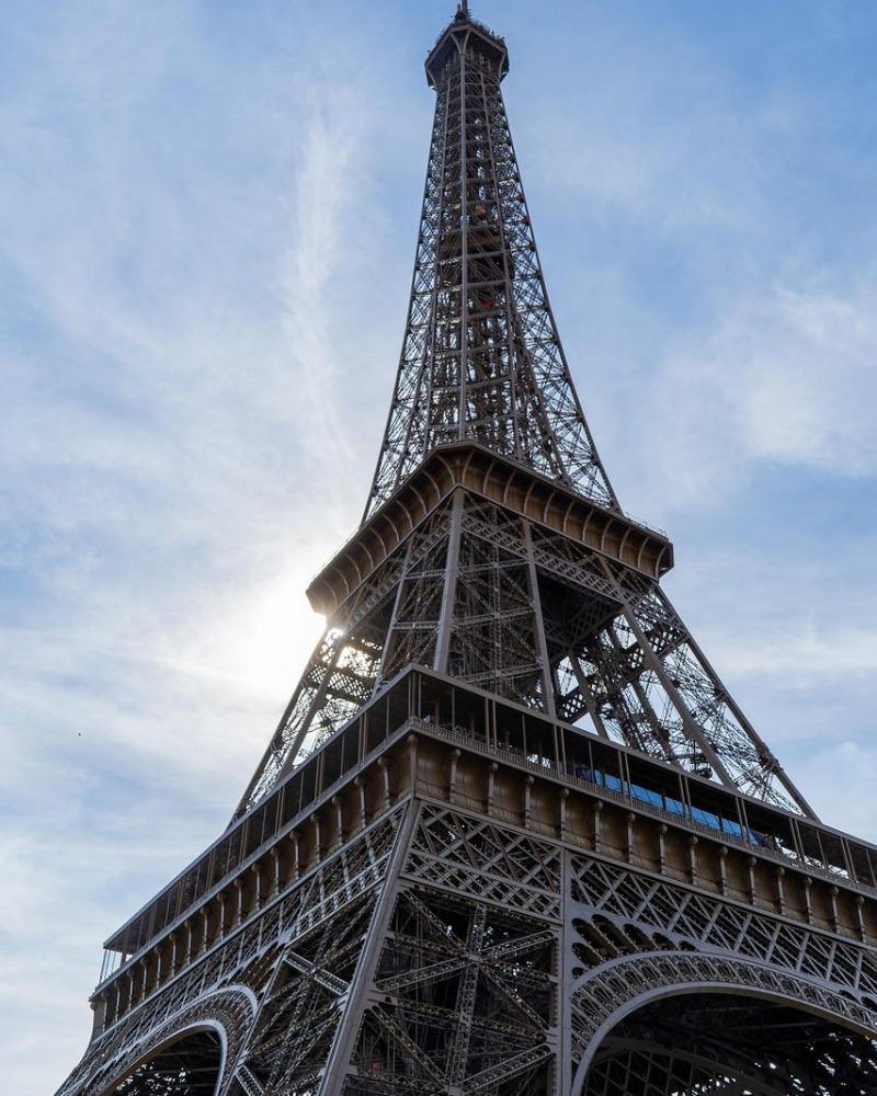 Low-angle view of the Eiffel Tower against a partly cloudy blue sky, with sunlight streaming through the structure.