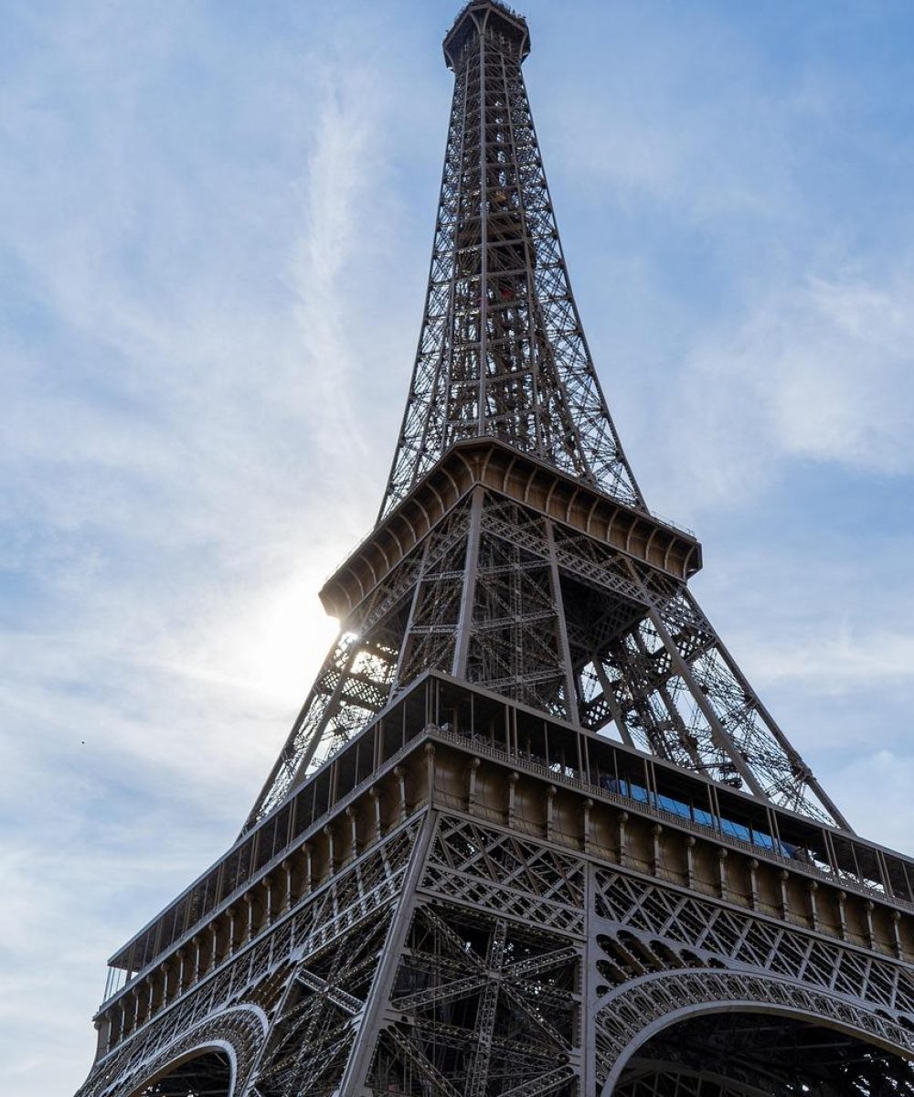 Vue en contre-plongée de la Tour Eiffel dans un ciel bleu partiellement nuageux, la lumière du soleil traversant la structure.