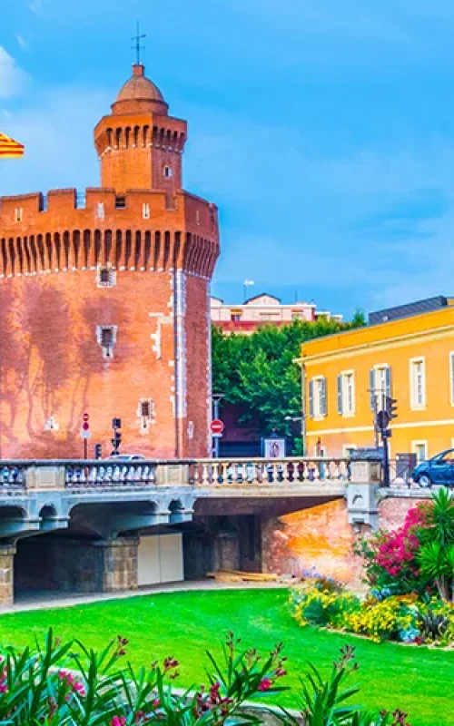 Une tour historique en briques rouges avec un drapeau se dresse près de bâtiments colorés et d'un pont, entourés de jardins verdoyants et de fleurs épanouies sous un ciel bleu lumineux.
