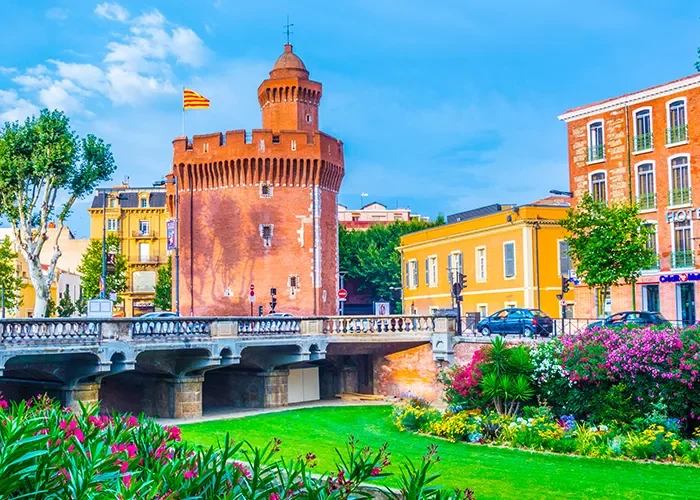 Une tour historique en briques rouges avec un drapeau se dresse près de bâtiments colorés et d'un pont, entourés de jardins verdoyants et de fleurs épanouies sous un ciel bleu lumineux.