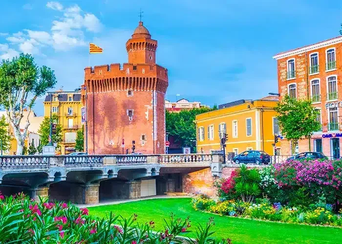 A historic red brick tower with a flag stands next to colourful buildings and a bridge, surrounded by lush gardens and flowers blooming under a bright blue sky.