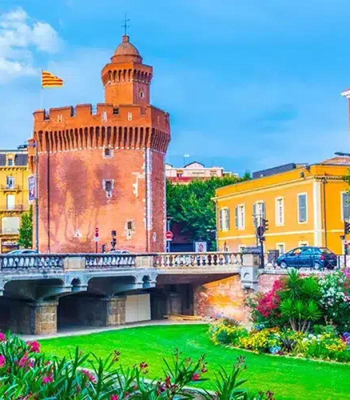 Une tour historique en briques rouges avec un drapeau se dresse près de bâtiments colorés et d'un pont, entourés de jardins verdoyants et de fleurs épanouies sous un ciel bleu lumineux.