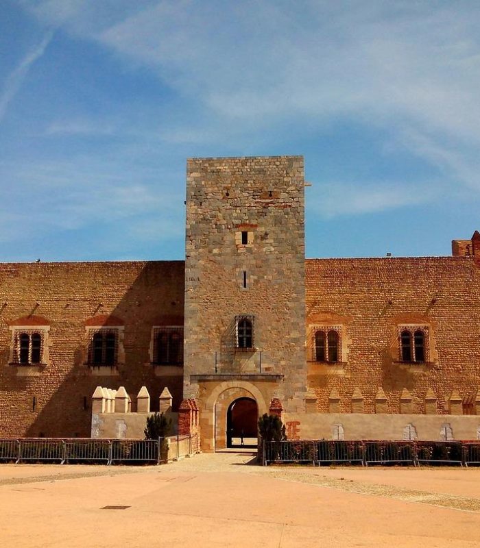 A large rectangular stone fortress, with a square central tower and vaulted entrance, stands under a blue sky, surrounded by a vast open courtyard and low barricades.