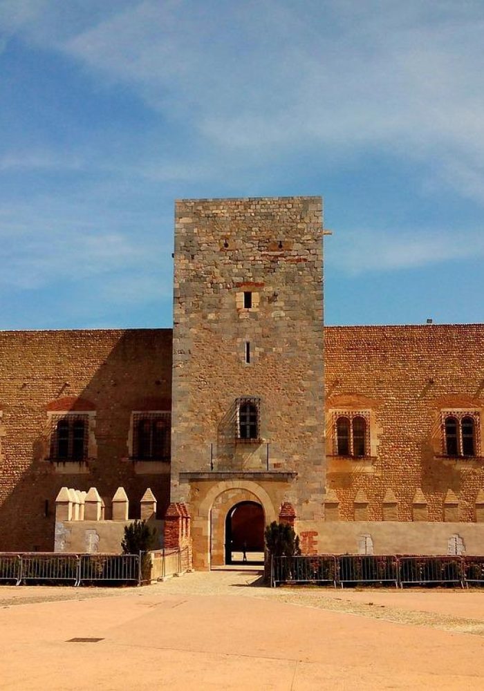 Une grande forteresse rectangulaire en pierre, avec une tour centrale carrée et une entrée voûtée, se dresse sous un ciel bleu, entourée d'une vaste cour ouverte et de barricades basses.