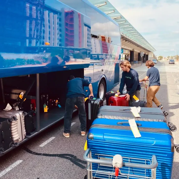 People load suitcases and luggage into a blue tour bus parked at a curb. Several large suitcases, ready for consigne bagages France or transport bagages Europe, are on a luggage cart in the sunny foreground.