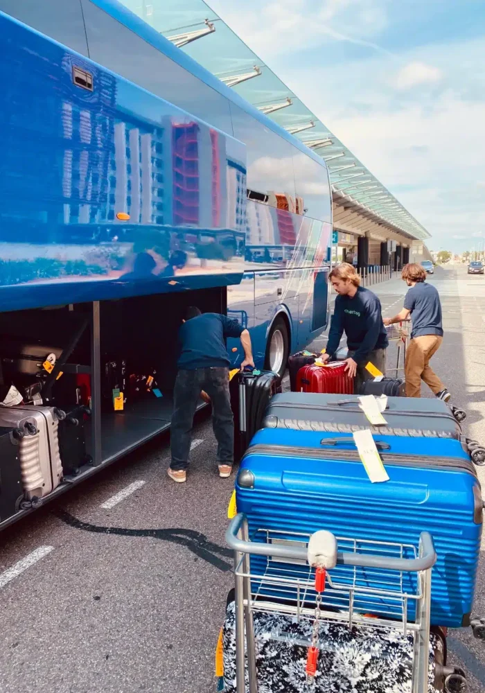 People load suitcases and luggage into a blue tour bus parked at a curb. Several large suitcases, ready for consigne bagages France or transport bagages Europe, are on a luggage cart in the sunny foreground.