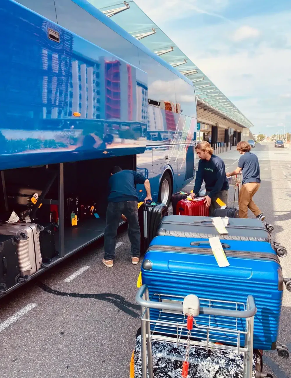 People load suitcases and luggage into a blue tour bus parked at a curb. Several large suitcases, ready for consigne bagages France or transport bagages Europe, are on a luggage cart in the sunny foreground.