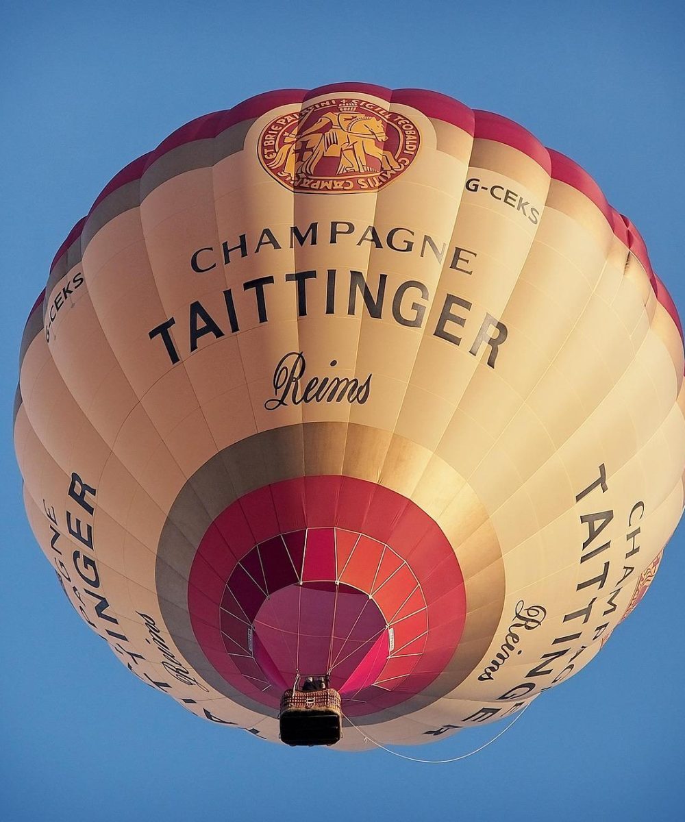 Une montgolfière portant la marque "Champagne Taittinger Reims" flotte dans un ciel bleu clair, vue d'en bas. La montgolfière est de couleur crème avec des accents rouges et or et un panier en osier visible.