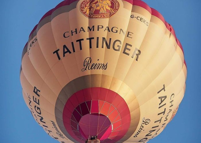 A hot air balloon bearing the brand name "Champagne Taittinger Reims" floats in a light blue sky, seen from below. The balloon is cream-coloured with red and gold accents and a visible wicker basket.