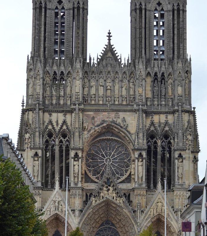 A large Gothic cathedral with two tall towers, intricate stone carvings, pointed arches and a large circular stained glass rose window above the main entrance. Trees line the street in front of the cathedral.