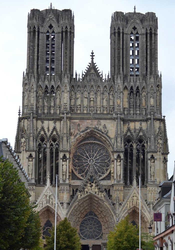 Une grande cathédrale gothique avec deux hautes tours, des sculptures de pierre complexes, des arcs en ogive et une grande rosace circulaire en vitrail au-dessus de l'entrée principale. Des arbres bordent la rue devant la cathédrale.