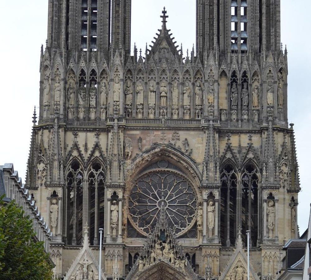 Une grande cathédrale gothique avec deux hautes tours, des sculptures de pierre complexes, des arcs en ogive et une grande rosace circulaire en vitrail au-dessus de l'entrée principale. Des arbres bordent la rue devant la cathédrale.