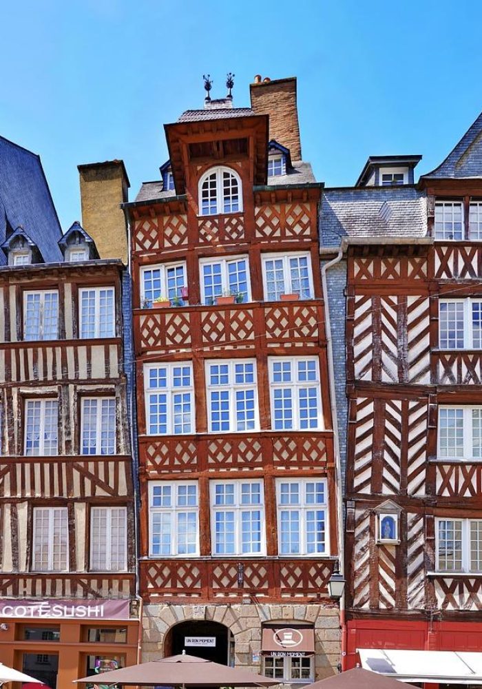 Colourful half-timbered buildings with ornate wooden motifs line a street under a bright blue sky; outdoor café tables with red umbrellas can be seen in the foreground.