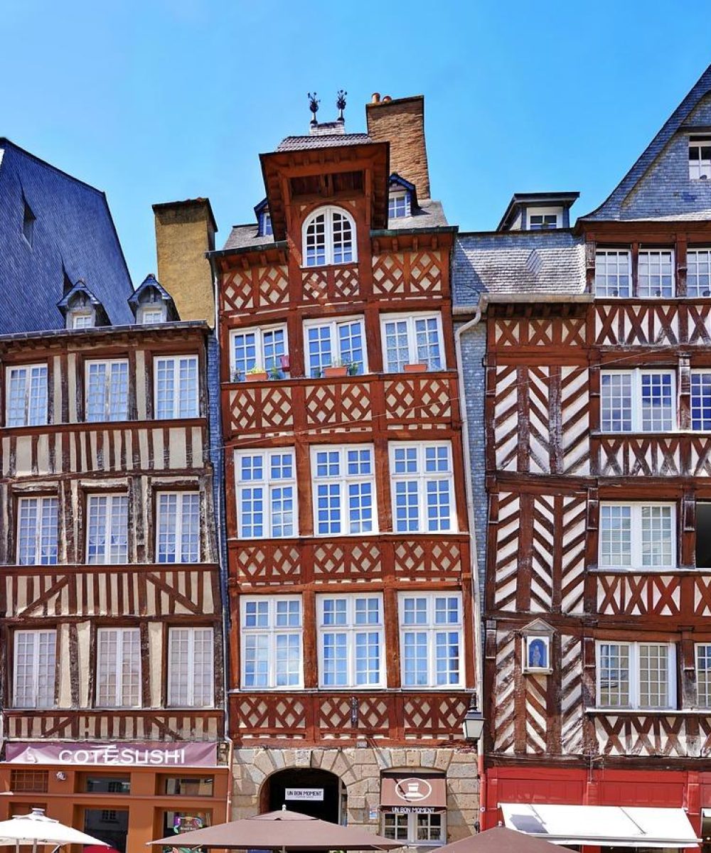 Des bâtiments à colombages colorés avec des motifs en bois ornés bordent une rue sous un ciel bleu vif ; des tables de café en plein air avec des parapluies rouges sont visibles au premier plan.