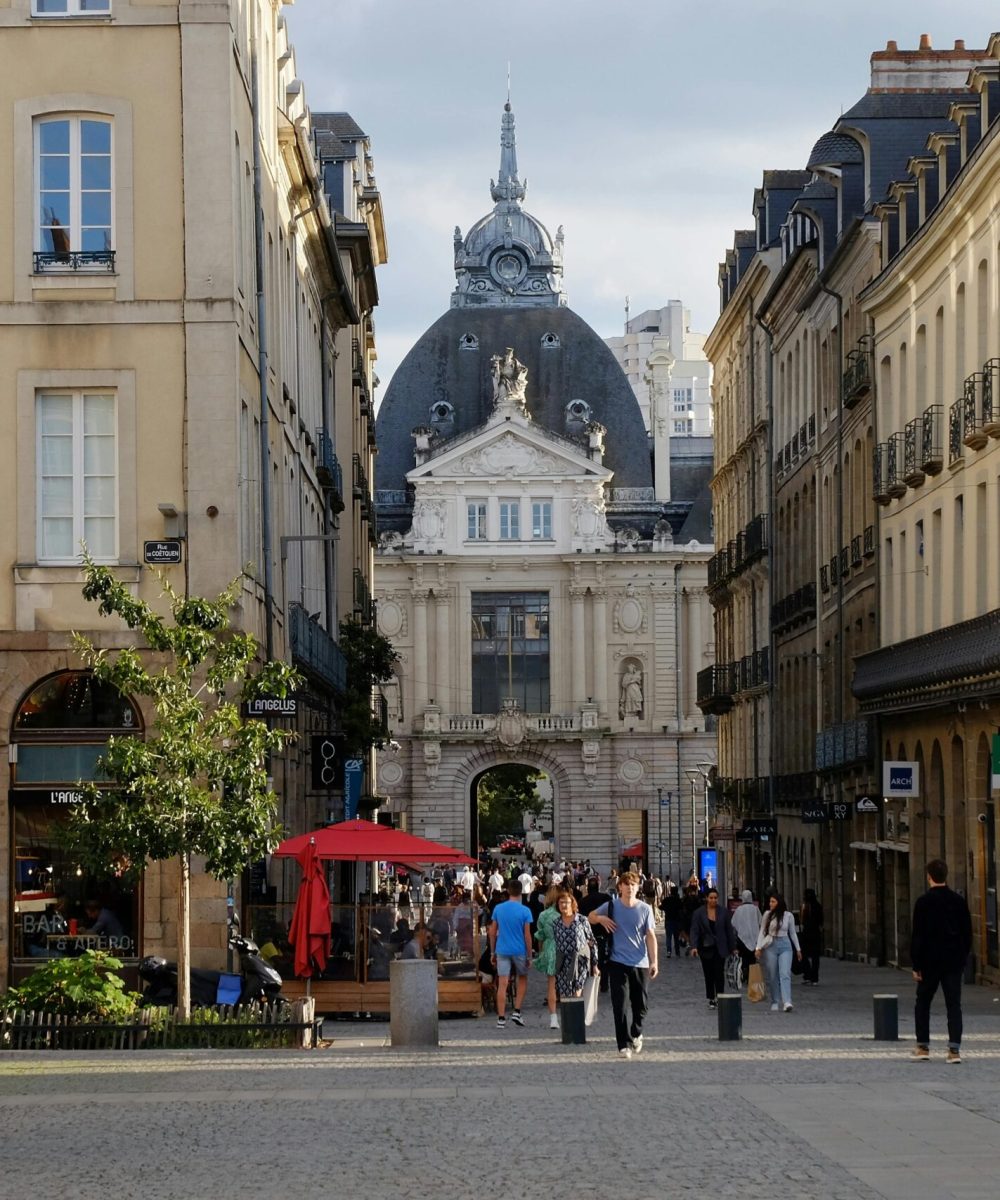 Une scène de rue animée dans une ville européenne avec des gens qui marchent, des sièges en plein air, des bâtiments historiques en pierre et un grand bâtiment en forme de dôme à l'arrière-plan sous un ciel nuageux.