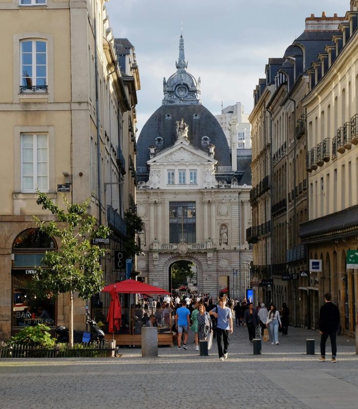 A lively street scene in a European city with people walking, outdoor seating, historic stone buildings and a large domed building in the background under a cloudy sky.