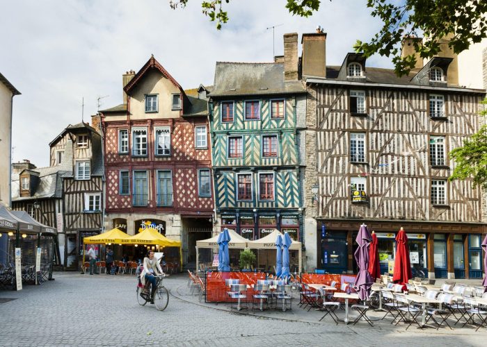 Un cycliste traverse une place pavée bordée de bâtiments à colombages colorés, de cafés en plein air et de tables vides avec des parapluies fermés dans une ville européenne historique.