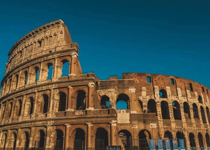 Le Colisée de Rome, en Italie, est représenté sous un ciel bleu clair. Les arches de l'ancien amphithéâtre en pierre et l'extérieur usé par les intempéries sont bien visibles, baignés par la lumière chaude du soleil.