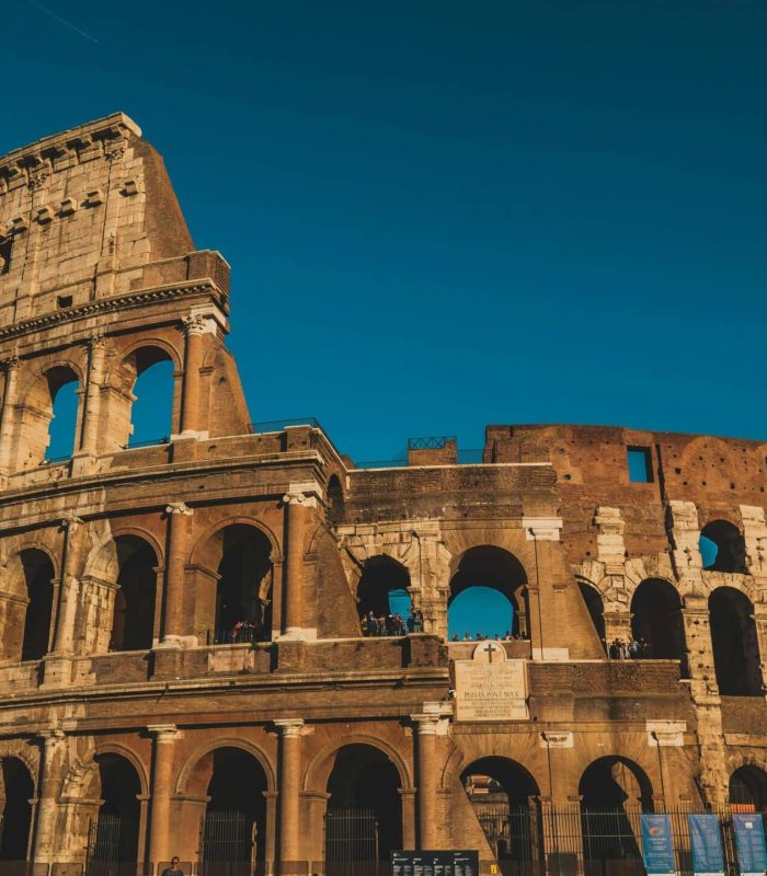 Le Colisée de Rome, en Italie, est représenté sous un ciel bleu clair. Les arches de l'ancien amphithéâtre en pierre et l'extérieur usé par les intempéries sont bien visibles, baignés par la lumière chaude du soleil.
