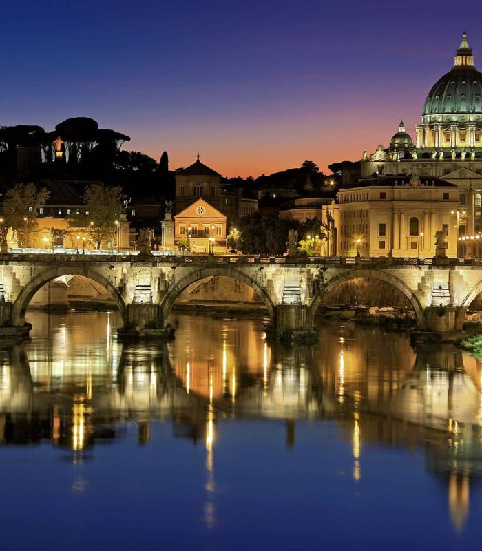 Vue nocturne de la basilique Saint-Pierre et du Vatican à Rome, illuminés, se reflétant dans le Tibre, avec le pont historique Ponte Sant'Angelo au premier plan.