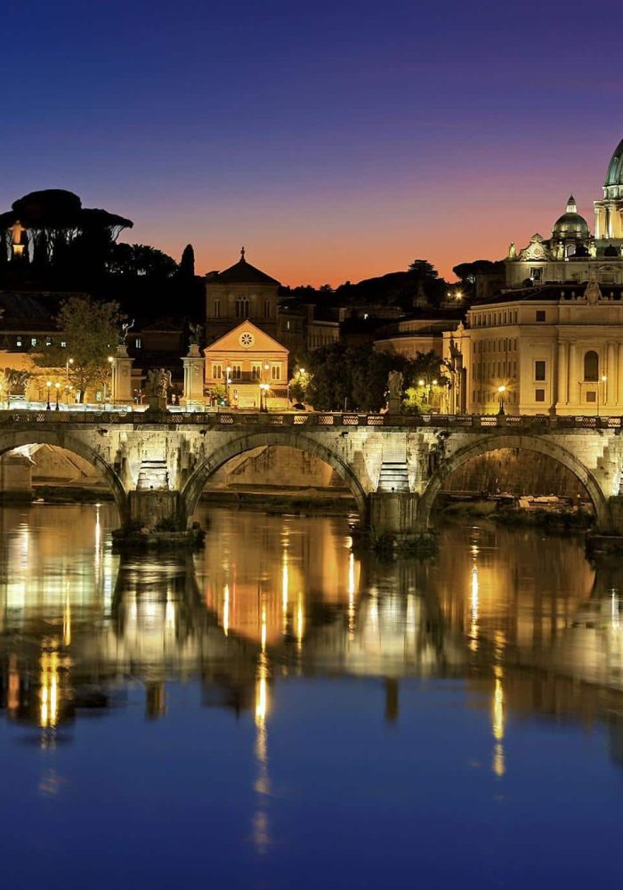 Vue nocturne de la basilique Saint-Pierre et du Vatican à Rome, illuminés, se reflétant dans le Tibre, avec le pont historique Ponte Sant'Angelo au premier plan.