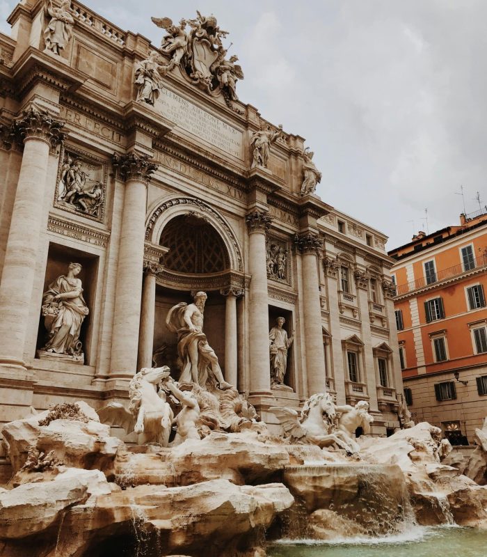 L'image montre la fontaine de Trevi à Rome, en Italie, une fontaine baroque ornée de statues de personnages mythologiques, de l'eau s'écoulant dans un grand bassin et des bâtiments historiques en arrière-plan, sous un ciel nuageux.