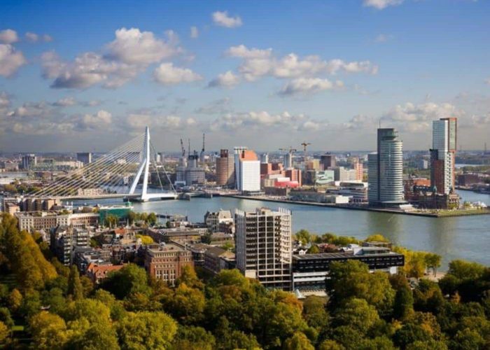 Paysage urbain de Rotterdam avec le pont Erasmus qui enjambe le fleuve, entouré de gratte-ciel modernes, d'eau et d'arbres verts sous un ciel bleu avec des nuages épars.