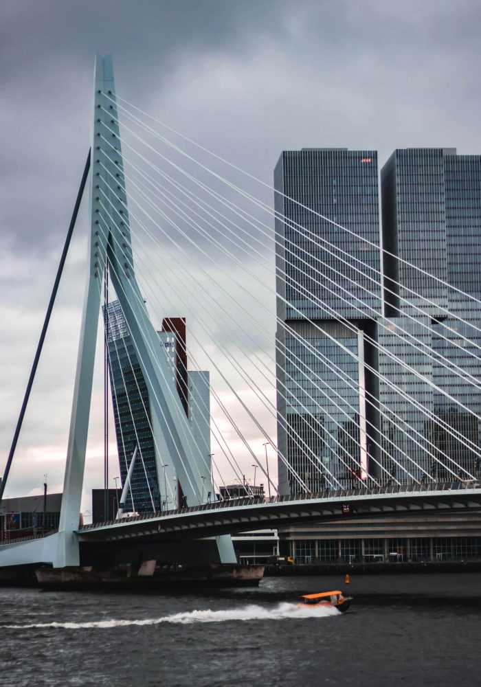 Un pont moderne à haubans enjambe une rivière et un bateau rapide passe en dessous, avec en toile de fond de grands gratte-ciel en verre sous un ciel nuageux.
