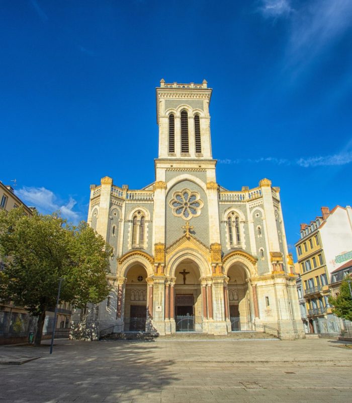 Une grande église en pierre avec une haute tour centrale et trois entrées voûtées se dresse sur une place ouverte et ensoleillée, entourée d'immeubles résidentiels, sous un ciel d'un bleu éclatant.