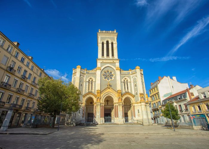 A large stone church with a high central tower and three vaulted entrances stands in an open, sunny square, surrounded by residential buildings, under a brilliant blue sky.