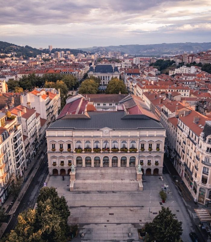 Aerial view of a large historic building with a square in front, surrounded by red-roofed buildings in a city at sunset, with hills and a partly cloudy sky in the background.
