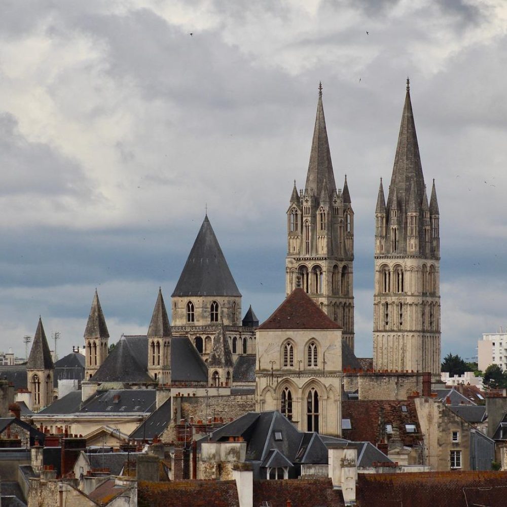 Un paysage urbain avec des bâtiments historiques en pierre et des tours d'église à pointes multiples sous un ciel nuageux, avec des bâtiments modernes visibles à l'arrière-plan.