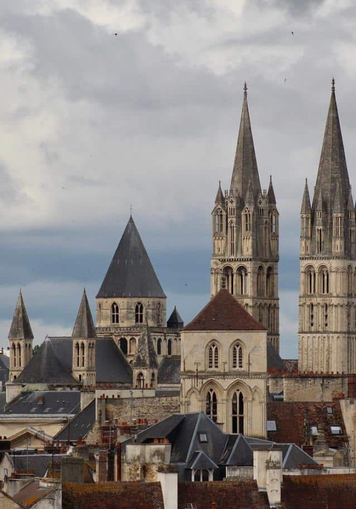 A cityscape of historic stone buildings and multi-pointed church towers under a cloudy sky, with modern buildings visible in the background.