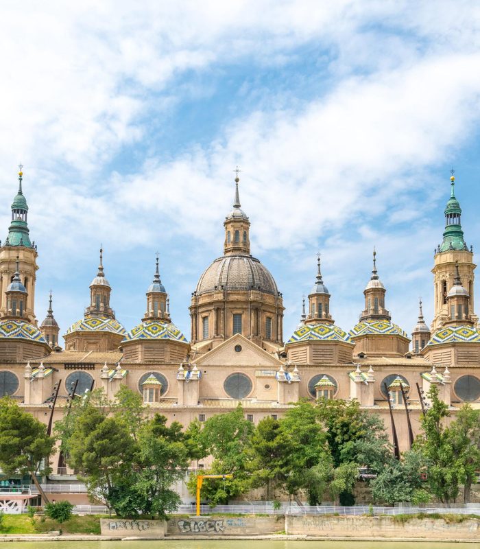 Une grande cathédrale ornée de multiples dômes et de hautes tours, aux détails complexes et aux toits de tuiles colorées, se dresse derrière des arbres sous un ciel partiellement nuageux.