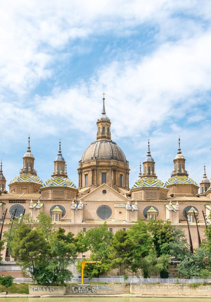 Une grande cathédrale ornée de multiples dômes et de hautes tours, aux détails complexes et aux toits de tuiles colorées, se dresse derrière des arbres sous un ciel partiellement nuageux.