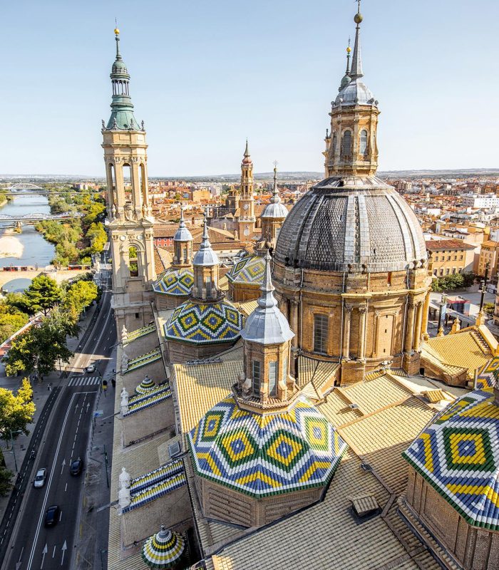 Vue aérienne de la Basílica de Nuestra Señora del Pilar avec ses dômes en tuiles colorées, située au bord de l'Ebre à Saragosse, Espagne, avec la ville et un pont en pierre en arrière-plan lors d'une journée ensoleillée.