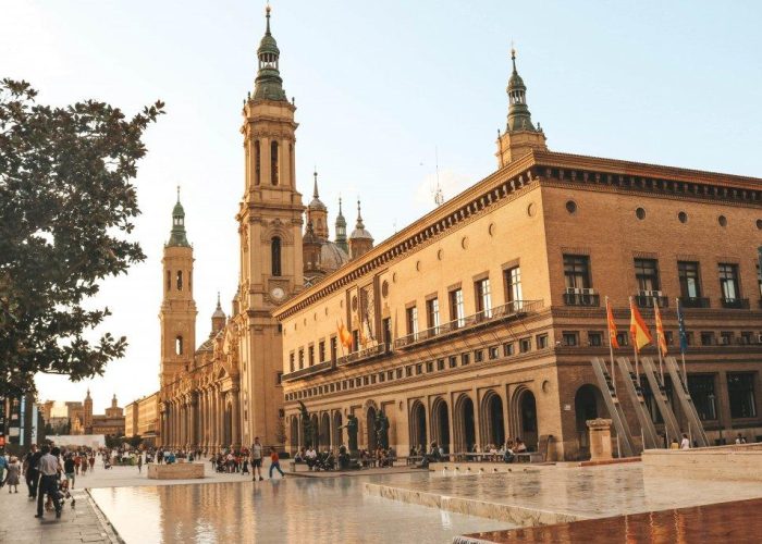 Des personnes marchent près d'un grand bâtiment historique aux fenêtres cintrées et aux hautes tours, à côté d'une place avec un bassin réfléchissant et des drapeaux, sous un ciel clair au coucher du soleil.