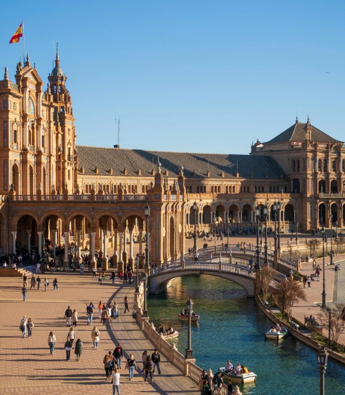 Les gens marchent et rament le long du canal de la Plaza de España à Séville, en Espagne, le drapeau espagnol flottant au sommet des bâtiments ornés et illuminés par le soleil, sous un ciel bleu limpide.