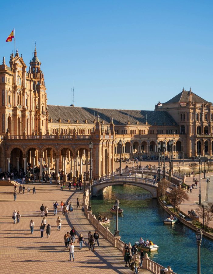 Les gens marchent et rament le long du canal de la Plaza de España à Séville, en Espagne, le drapeau espagnol flottant au sommet des bâtiments ornés et illuminés par le soleil, sous un ciel bleu limpide.