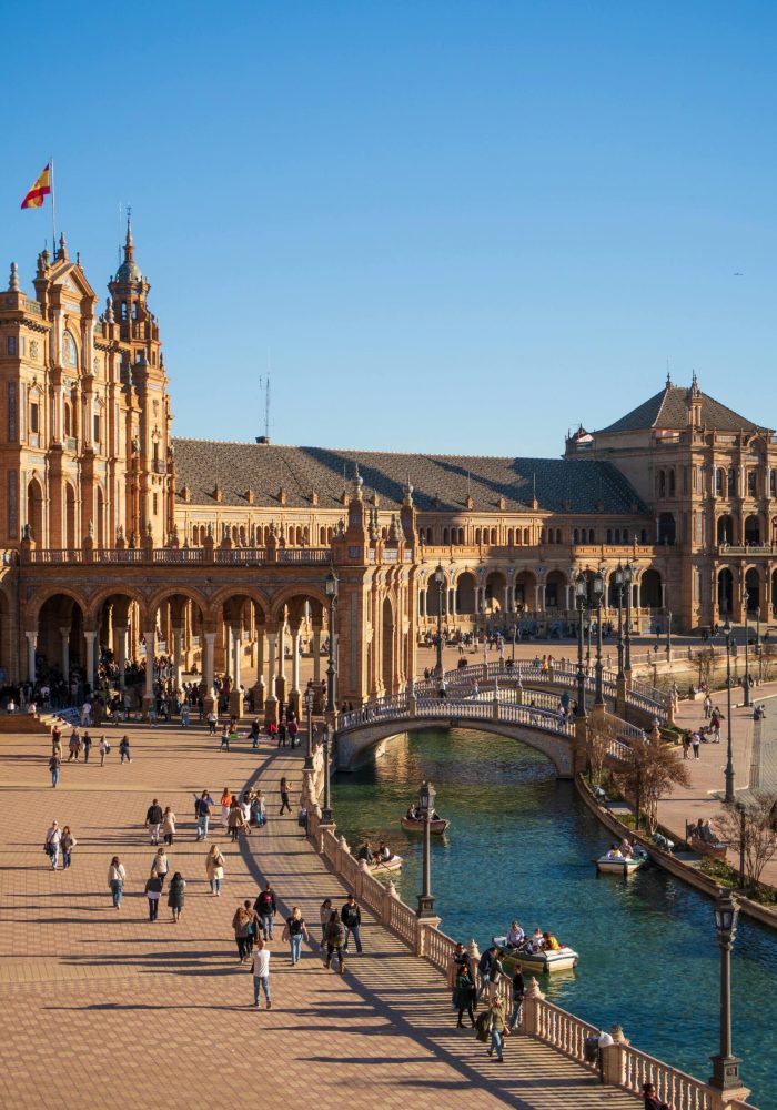 Les gens marchent et rament le long du canal de la Plaza de España à Séville, en Espagne, le drapeau espagnol flottant au sommet des bâtiments ornés et illuminés par le soleil, sous un ciel bleu limpide.