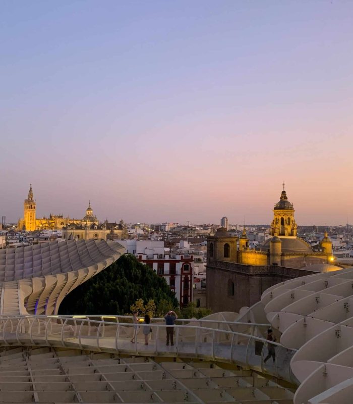 Vue de Séville au coucher du soleil, avec la structure moderne Metropol Parasol au premier plan et les tours des églises historiques et les bâtiments de la ville au loin, sous un ciel clair.