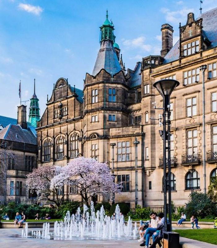Des personnes se détendent près d'une petite fontaine et d'un cerisier en fleurs sur une place de la ville entourée d'un bâtiment historique orné, sous un ciel bleu.