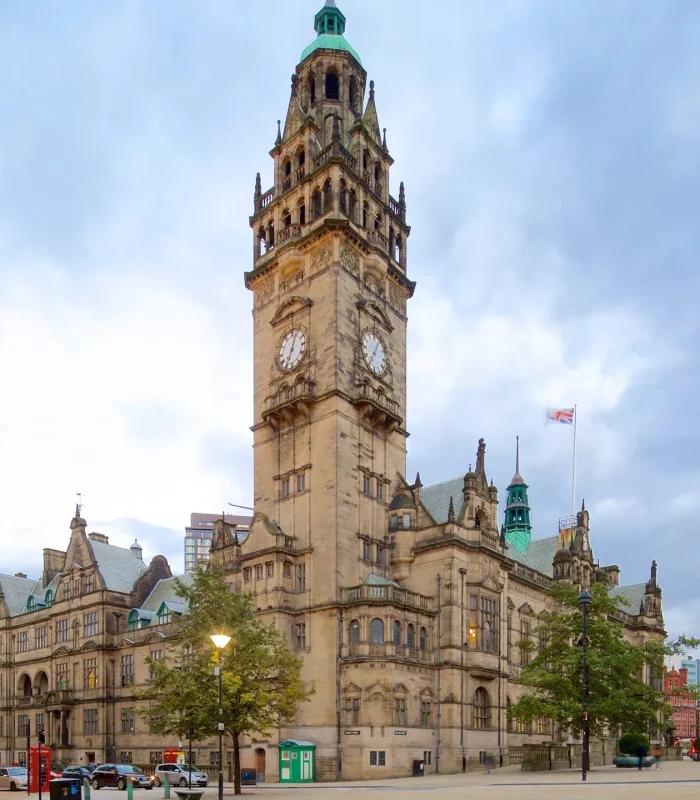 Un grand bâtiment historique avec une haute tour d'horloge se dresse sur une place de la ville, entouré d'arbres et de bâtiments voisins sous un ciel nuageux. Des personnes et des voitures sont visibles au premier plan.