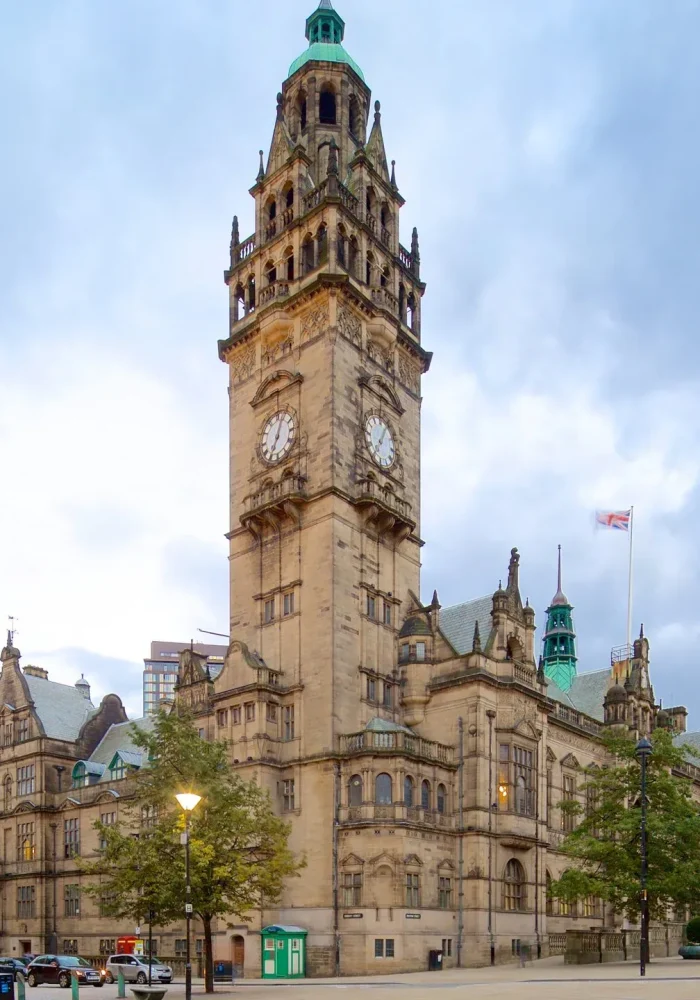 Un grand bâtiment historique avec une haute tour d'horloge se dresse sur une place de la ville, entouré d'arbres et de bâtiments voisins sous un ciel nuageux. Des personnes et des voitures sont visibles au premier plan.