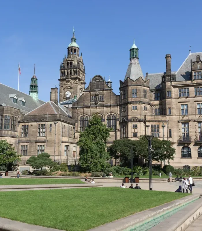 Un grand bâtiment historique avec une architecture en pierre ornée, des tours et une tour d'horloge, entouré d'une cour paysagée avec des pelouses vertes, des arbres et des gens qui se détendent sous un ciel bleu clair.