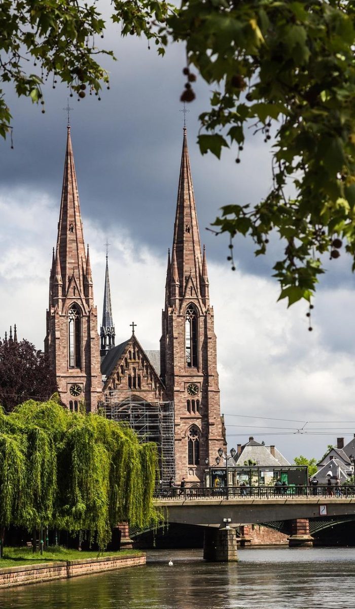 Une grande église gothique aux deux flèches pointues se dresse au bord d'une rivière, partiellement encadrée par des arbres verts et des branches en surplomb, avec un pont devant et des nuages sombres dans le ciel.