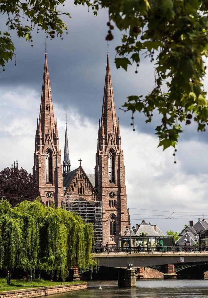 A large Gothic church with two pointed spires stands on the banks of a river, partly framed by green trees and overhanging branches, with a bridge in front and dark clouds in the sky.