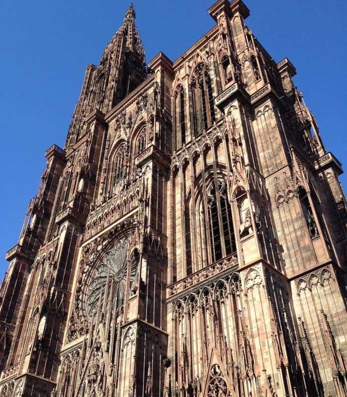 Vue en contre-plongée de la cathédrale de Strasbourg, avec son architecture gothique complexe, ses arcs brisés et ses sculptures en pierre détaillées, dans un ciel bleu clair.